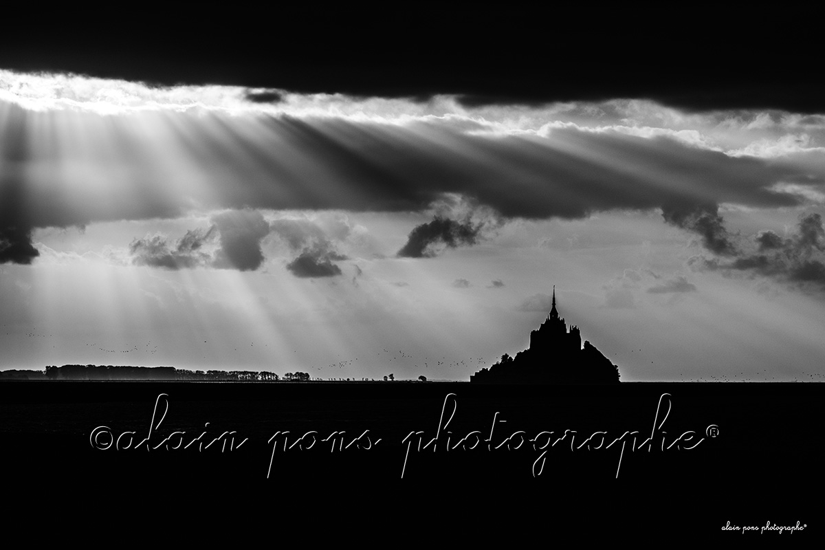 Photographie en noir et blanc du Mont-Saint-Michel sous des rayons lumineux,