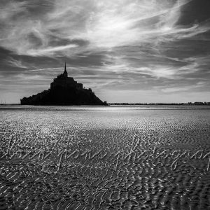 Photographie du Mont-Saint-Michel avec le sable de l'estran en premier plan à marée basse, en noir et blanc.