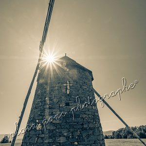 Photographie du moulin à vent à paroles sur le plateau d'Ally en Haute-Loire, format vertical, en noir et blanc sépia, et le soleil en étoile sur le toit du moulin.